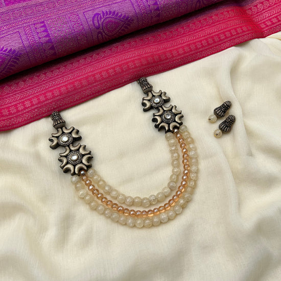 A beige-colored terracotta beaded neckwear with studs, displayed on a white fabric. The neckwear features a series of round and oval-shaped beads, with a central decorative stud.