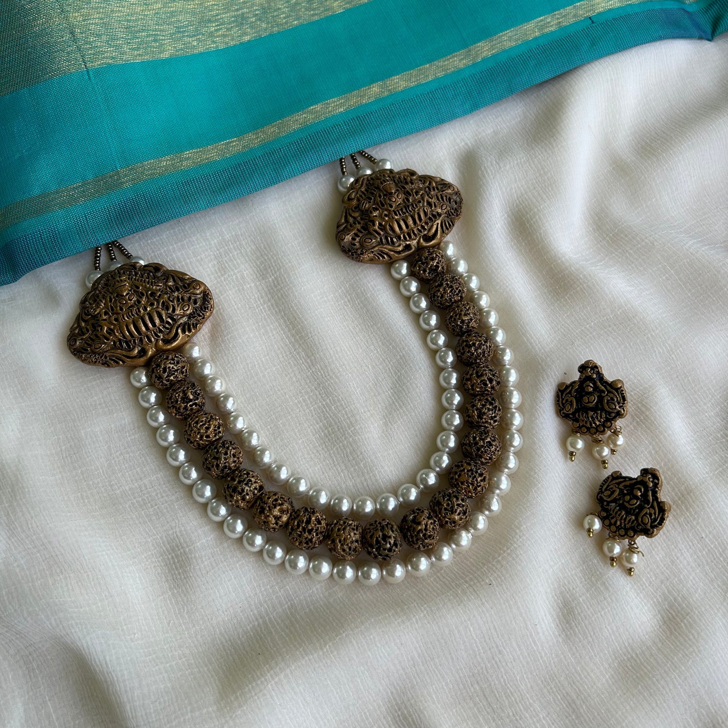 A set of handcrafted terracotta jewelry including a beaded necklace with gold shades and studs, and matching earrings, displayed on a white fabric backdrop.